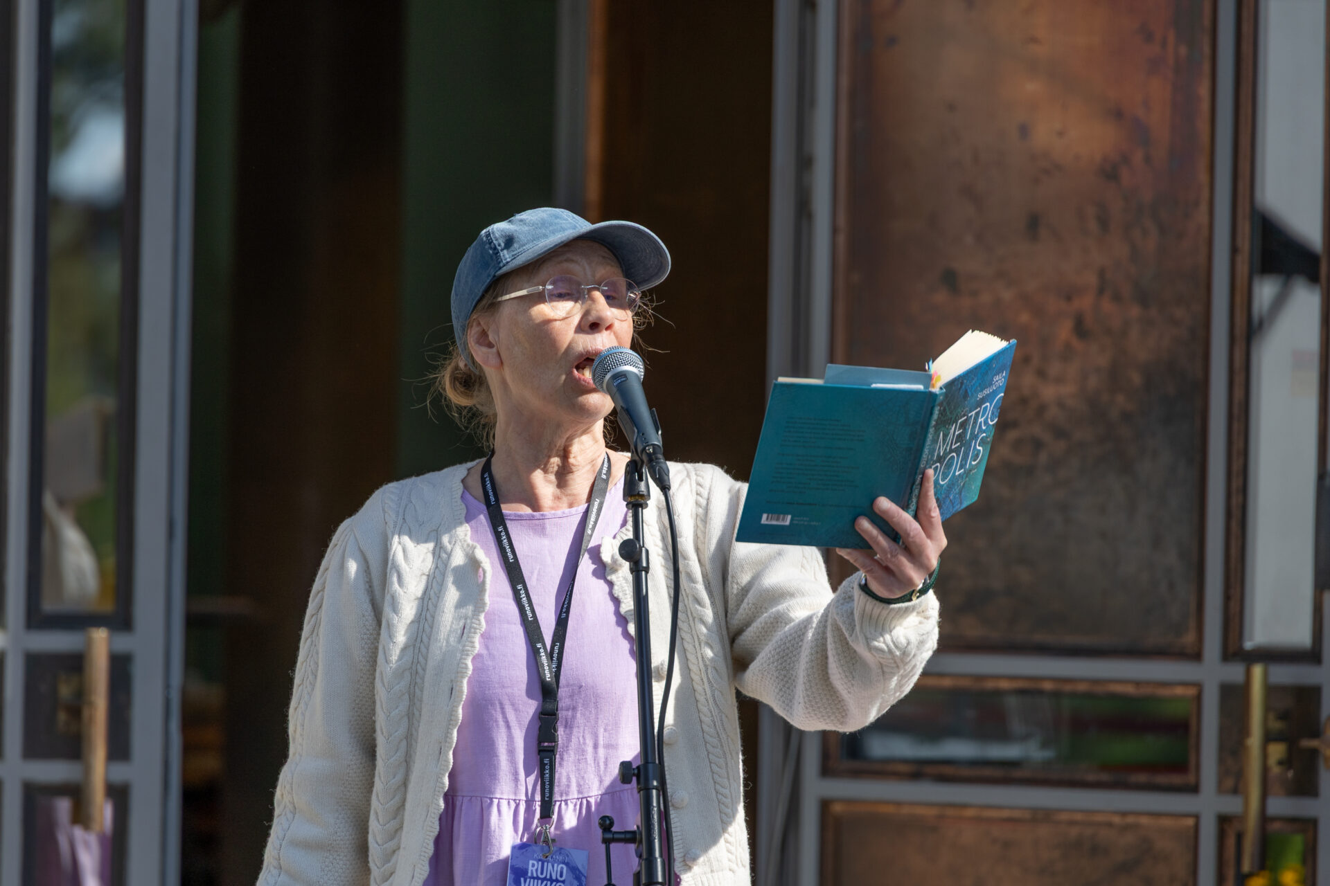 Kati Outinen pitää kädessään kirjaa ja puhuu mikrofoniin. Päällään valkoinen neuletakki, lila paita ja sininen lippalakki. Kaulassa Runoviikon henkilökortti.