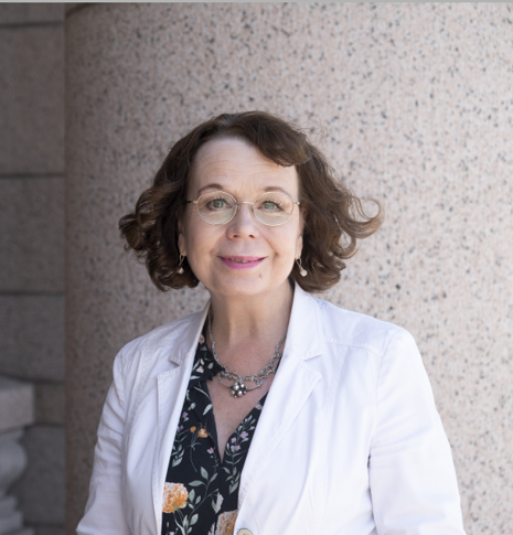 Kiviseinää vasten otettu kuva, jossa Pia Viitanen puolivartalokuvassa. Ruskeat puolipitkän hiukset kiharoina. Kasvoilla pieni hymy. Valkoinen jakku ja kukallinen paita sen alla.